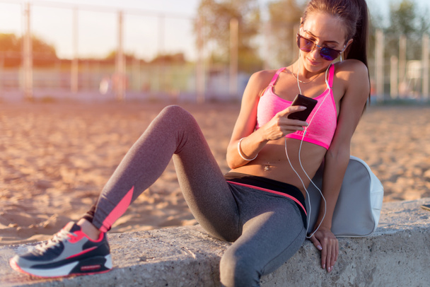 Beautiful fitness athlete woman wearing sunglasses resting listening music after work out exercising on summer evening in beach at sunset with smartphone outdoor portrait. - Fotografie, Obrázek