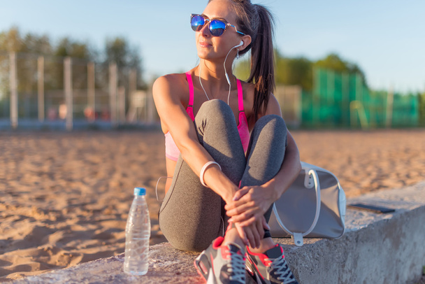 Hermosa atleta de fitness mujer con gafas de sol escuchando música descansando después de hacer ejercicio en la noche de verano en la playa al atardecer retrato al aire libre
. - Foto, Imagen