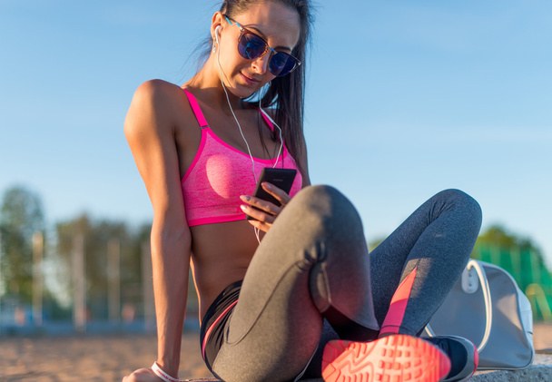 Gyönyörű fitness sportoló nő pihenő hallgat zenét, gyakorlása a nyári este Beach napnyugtakor smartphone szabadtéri portré munka után napszemüveget visel. - Fotó, kép