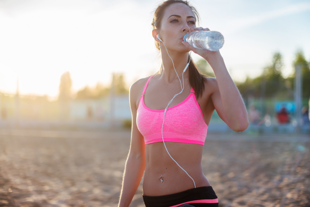 Γυναίκα όμορφη καταλληλότητας αθλητής που αναπαύεται πόσιμο νερό μετά τη δουλειά έξω άσκηση στην παραλία το βράδυ καλοκαίρι σε ηλιόλουστες ηλιοφάνειας υπαίθρια πορτρέτο - Φωτογραφία, εικόνα