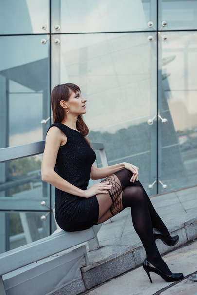 Fashion outdoor portrait of sexy beautiful woman with brown hair wearing in black dress, hose and high heels. Posing on creative urban background. - Photo, Image