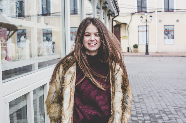 young hipster very beautiful caucasian stilysh and trendy girl with gorgeous brunette hair is having fun smiling outdoors on a background of the streets and wooden cool backgrounds during windy spring day - Foto, Imagen