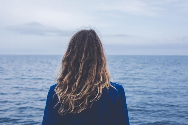 Młoda blondynka kręcone dziewczyna patrząc na mgliste słońca dzięki gęstej mgle na spokojne morze i błękitnym niebem widok z tyłu - Zdjęcie, obraz