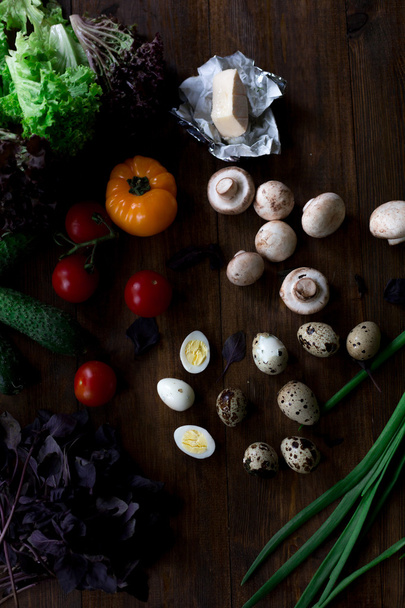 salad preparation with quail eggs, mushrooms, mix of red yellow and cherry tomatoes, cucumber, basil, brie cheese, lettuce, and black olives all organic and fresh from farmers market in rustic sryle on dark wood backround soft focus overhead-angle - Foto, Imagen