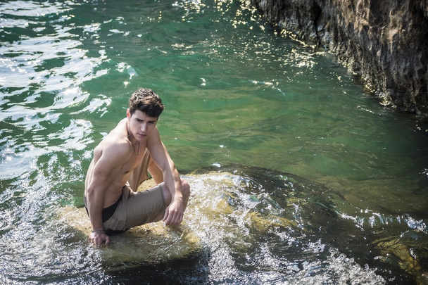 Молодий спортсмен без сорочки сидить на скелі біля моря
 - Фото, зображення