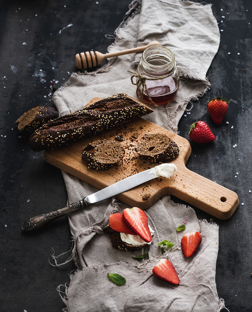 Breakfast set. Black baguette toasts with fresh strawberries, honey and mascarpone cheese on dark grunge table surface - Фото, изображение