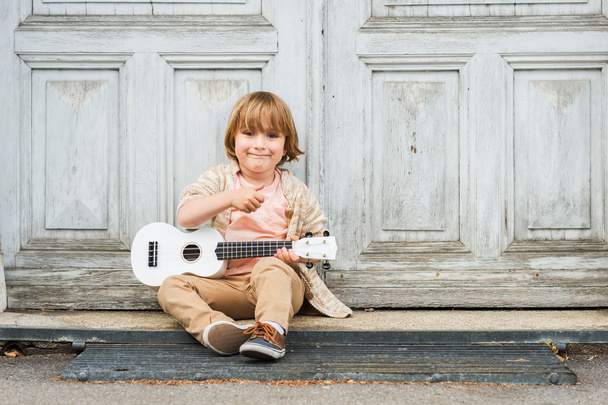 Little happy boy plays his guitar or ukulele, sitting by the wooden door outdoors - Photo, Image