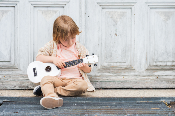 Petit garçon heureux joue de sa guitare ou ukulélé, assis près de la porte en bois à l'extérieur
 - Photo, image
