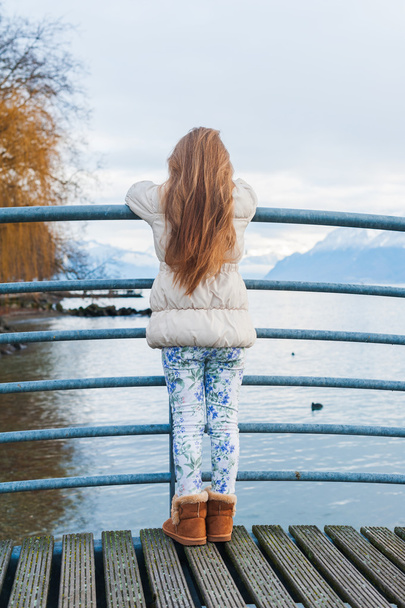 Cute little girl standing on a bridge, looking at the beautiful lake, wearing warm beige coat, back view - Fotoğraf, Görsel
