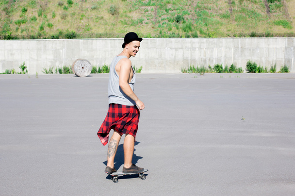 cool jonge en knappe Kaukasische brunette hipster skater jongen dragen van een hoed poseren glimlachend en plezier buiten terwijl het schaatsen met zijn skateboard tijdens geweldige zomerdag in de stad - Foto, afbeelding