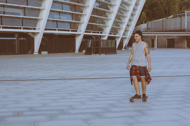 Cool mladý a pohledný kavkazské bruneta bederní bruslař chlap klobouk pózování s úsměvem a baví mimo při bruslení s jeho skateboard během úžasné letní den v městě - Fotografie, Obrázek