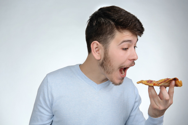 Young man eating piece of pizza on gray background - Φωτογραφία, εικόνα