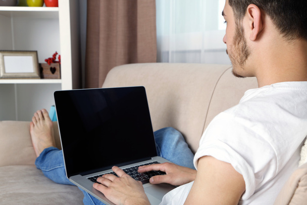 Красивий молодий чоловік сидить на дивані і використовує ноутбук в кімнаті
 - Фото, зображення