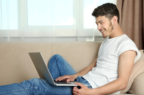 schöner junger Mann sitzt auf Sofa und benutzt Laptop im Zimmer - Foto, Bild