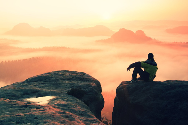 mounrains の谷の上のカラフルな夜明けを楽しみながら岩のピークの上に座って男性ハイカーの背面図 - 写真・画像