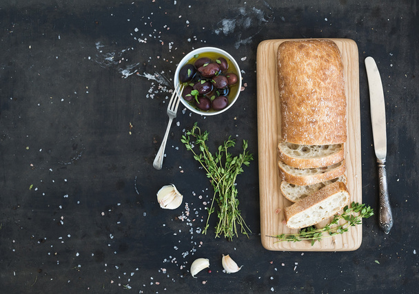 Italian ciabatta bread cut in slices on wooden chopping board with herbs, garlic and olives over dark grunge backdrop, copy space - Photo, image
