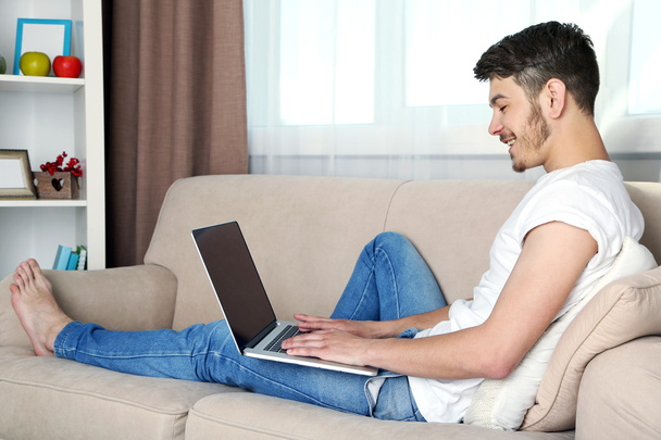Handsome young man sitting on sofa and using laptop in room - Fotografie, Obrázek