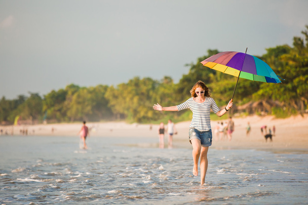 Cheerful young girl with rainbow umbrella having fun on the beach before sunset. Travel, holidays, vacation, healthy lifestyle concept - Фото, изображение