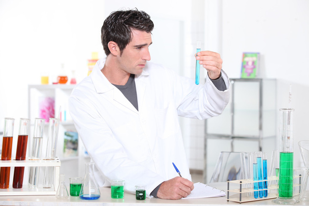 Young man studying the contents of a test tube and making notes accordingly - Photo, Image