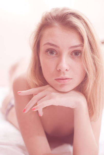 close up on sensually looking at camera naturally blond young pretty lady with silk skin, excellent hair & red nails having fun relaxing laying in bed on light copy space background portrait image - Foto, Imagen