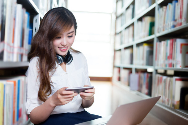 diáklány használ laptop és mozgatható telefon-ban könyvtár - Fotó, kép