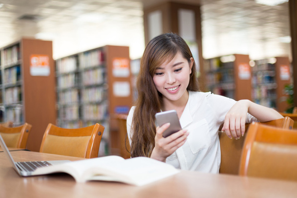 Φοιτήτριας χρησιμοποιώντας κινητό τηλέφωνο στη βιβλιοθήκη - Φωτογραφία, εικόνα