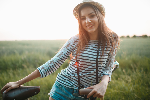 Adorável jovem mulher fica em um campo com sua bicicleta
 - Foto, Imagem