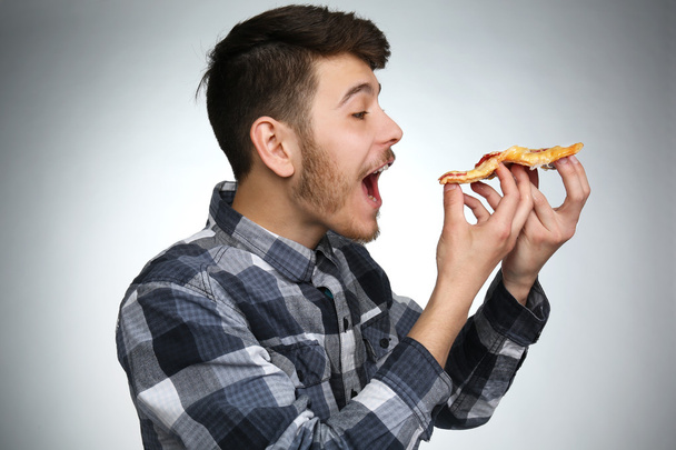 Young man eating piece of pizza on gray background - Фото, зображення