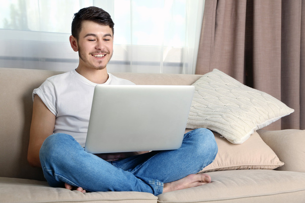 Handsome young man sitting on sofa and using laptop in room - Fotó, kép