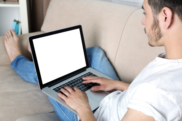 Красивый молодой человек сидит на диване и использует ноутбук в комнате
 - Фото, изображение