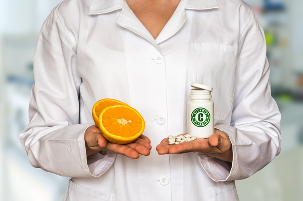 Young doctor holding two halves of orange and bottle of pills with vitamins and compare them - Fotoğraf, Görsel