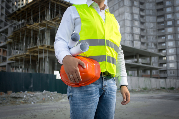Architect in jacket posing with red helmet at construction site - 写真・画像