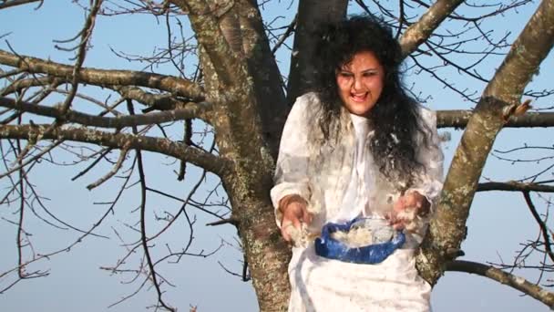 Verrückte dunkelhaarige Frau im langen weißen Nachthemd wirft Kissenfedern herum und sitzt auf einem Baum - Filmmaterial, Video