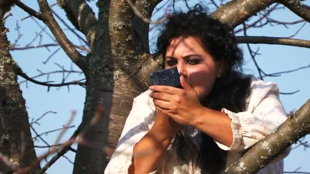 Verrückte dunkelhaarige Frau im langen weißen Nachthemd mit Lippenstift auf Baum sitzend - Filmmaterial, Video