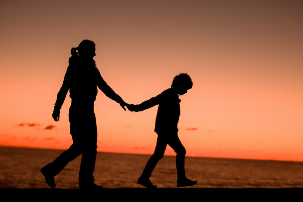 Silhouette of a young mother and her son walking along a pier in front of a sunset in the sky on a summer day - Zdjęcie, obraz