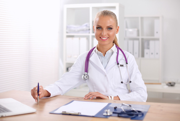 Όμορφη νεαρή χαμογελαστή γυναίκα γιατρός κάθεται στο γραφείο και γράφει. - Φωτογραφία, εικόνα