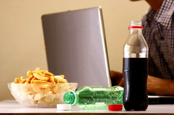 Пляшки безалкогольних напоїв або соди, чіпси і людина, що працює на ноутбуці на задньому плані
 - Фото, зображення