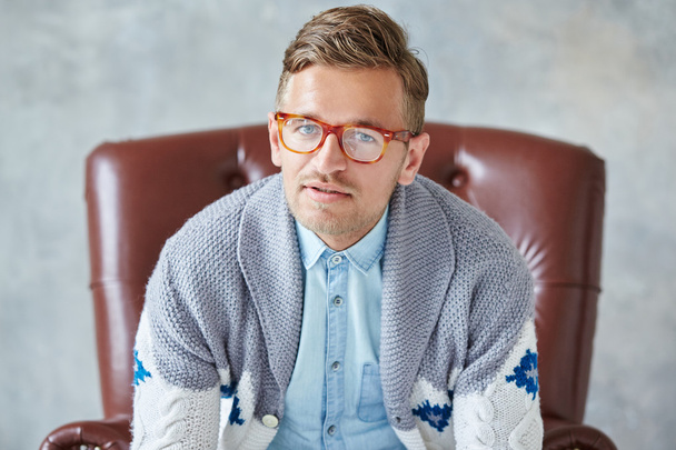 Portrait of a young intelligent man stares into the camera, good view, small unshaven, charismatic, wearing glasses rimmed with brown, blue shirt, gray sweater, sitting on a brown leather chair on a gray background - Fotoğraf, Görsel
