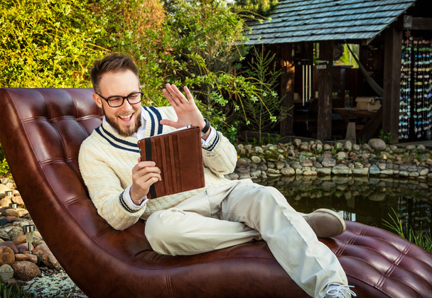 Young handsome man in glasses sit in luxury sofa with iPad in summer garden. - Foto, Imagen