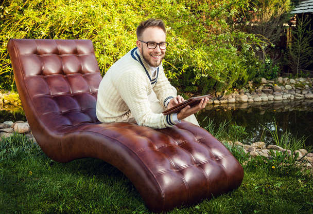 Young handsome man in glasses sit in luxury sofa with iPad in summer garden. - Foto, imagen