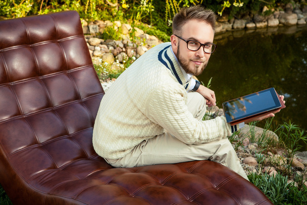 Young handsome man in glasses sit in luxury sofa with iPad in summer garden. - Photo, image