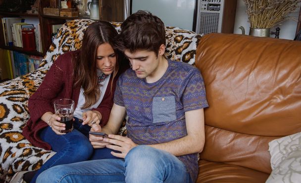 Joven pareja mirando teléfono inteligente sentado en el sofá
 - Foto, Imagen