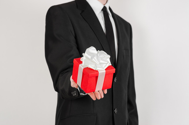 Theme holidays and gifts: a man in a black suit holds exclusive gift wrapped in red box with white ribbon and bow isolated on a white background in studio - Foto, Imagen