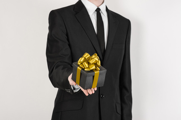 Theme holidays and gifts: a man in a black suit holds exclusive gift wrapped in a black box with gold ribbon and bow isolated on a white background in studio - Foto, Imagem