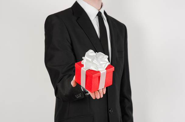 Theme holidays and gifts: a man in a black suit holds exclusive gift wrapped in red box with white ribbon and bow isolated on a white background in studio - Photo, image