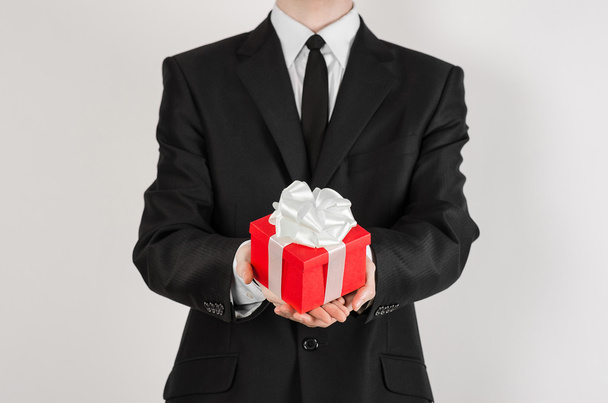 Theme holidays and gifts: a man in a black suit holds exclusive gift wrapped in red box with white ribbon and bow isolated on a white background in studio - Fotoğraf, Görsel