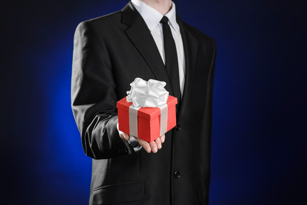 Theme holidays and gifts: a man in a black suit holds exclusive gift wrapped in red box with white ribbon and bow on a dark blue background in studio - Photo, image