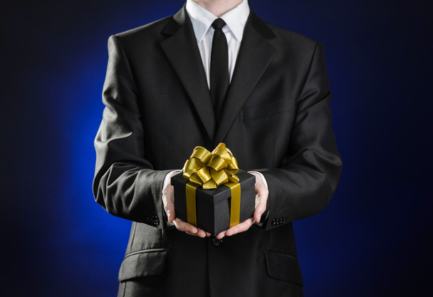 Theme holidays and gifts: a man in a black suit holds exclusive gift wrapped in a black box with gold ribbon and bow on a dark blue background in studio - Foto, Imagen