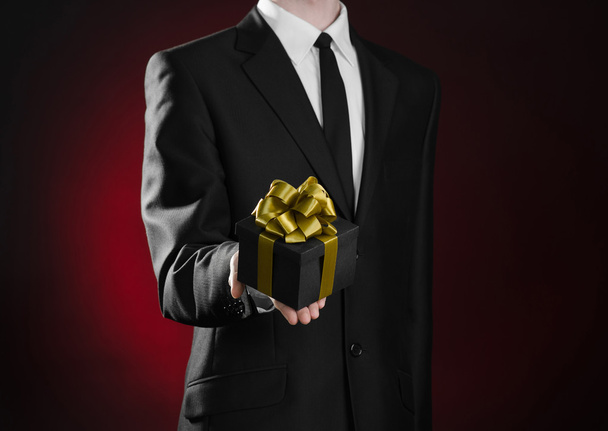 Theme holidays and gifts: a man in a black suit holds exclusive gift wrapped in a black box with gold ribbon and bow on a dark red background in studio - Fotografie, Obrázek