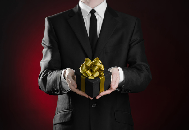 Theme holidays and gifts: a man in a black suit holds exclusive gift wrapped in a black box with gold ribbon and bow on a dark red background in studio - Valokuva, kuva